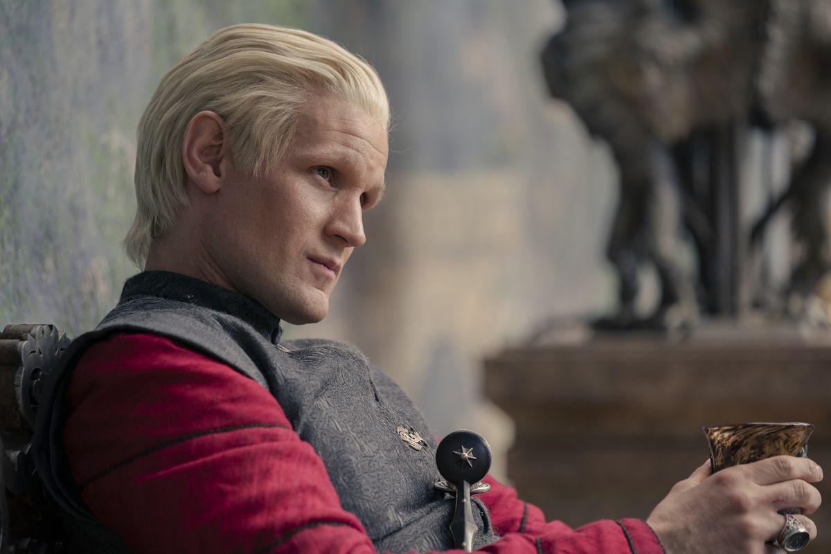 Daemon Targaryen, anointed, wearing a red shirt and gray robes, staring at someone while holding his wine in House of the Dragon