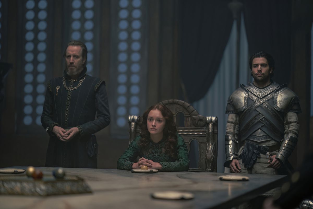 Alicent sits at the Small Council table with her hands folded on the table in front of her.  To her right stands her father with his hands slightly folded;  To her left, Ser Criston stands ready