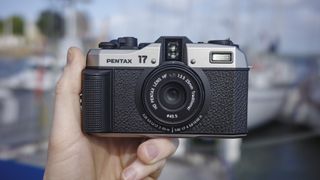 Pentax 17 compact film camera frontal, hand-held with boats in the background