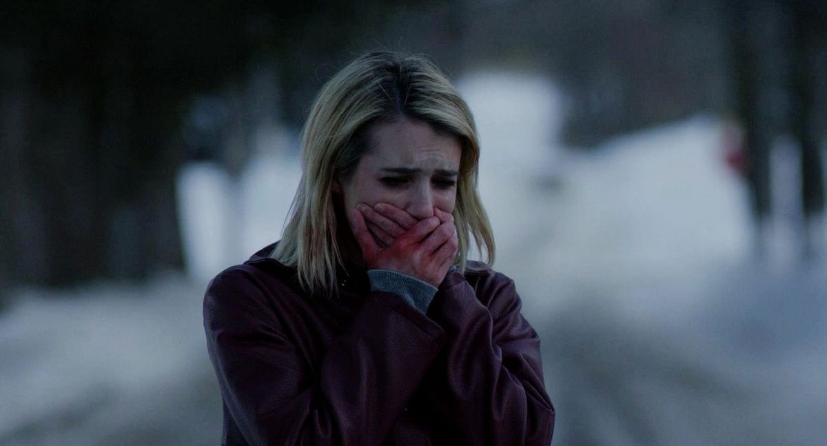 A woman covers her mouth with her hands, stained red, with a look of visible fear as she stares at something off-screen in The Blackcoat's Daughter.