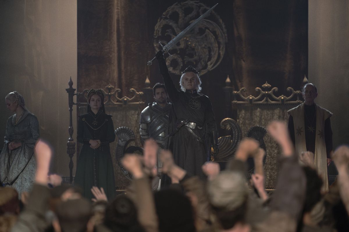 King Aegon II (Tom Glynn-Carney) stands before a crowd of cheering Westeros audience members and triumphantly holds his sword aloft