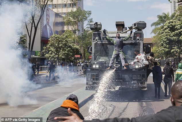 Protesters sit on a police water cannon as it tries to fend them off with chemically treated water jets next to an advertisement with a photo of Kenyan President William Ruto