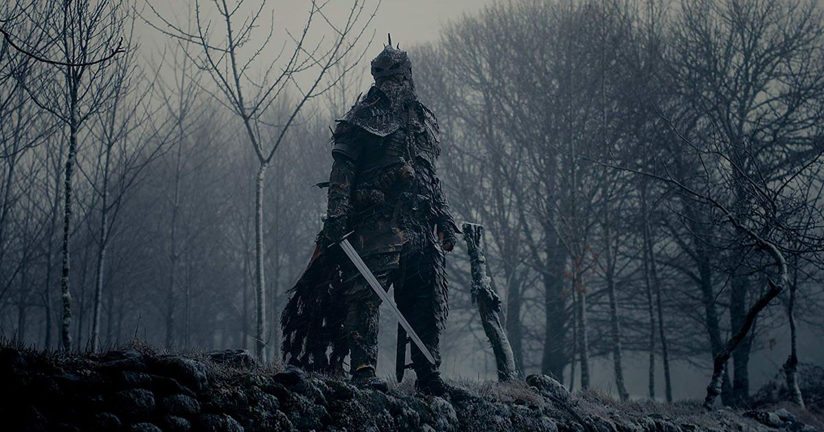 A warrior in black armor stands in front of a deserted winter forest.