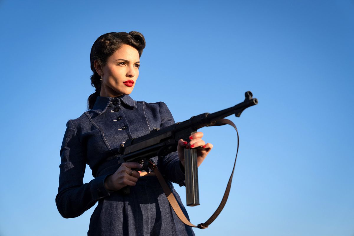 Eiza González, in a chic 1940s dress and deep red lipstick, holds a machine gun in The Ministry of Ungentlemanly Warfare