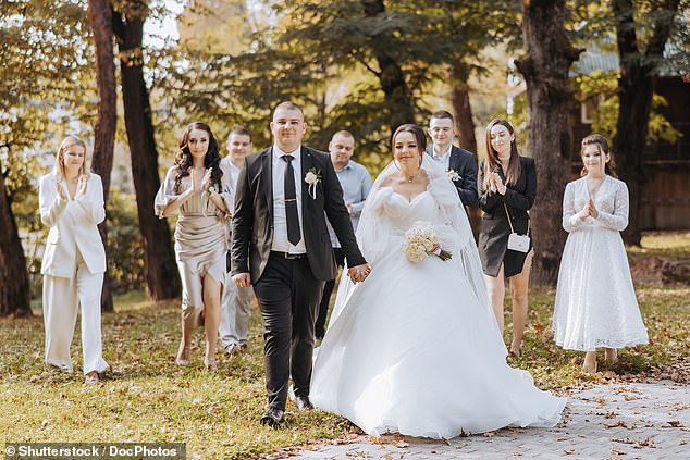 A bride and groom walk down a path with a group of people behind them, dressed in white (stock image)