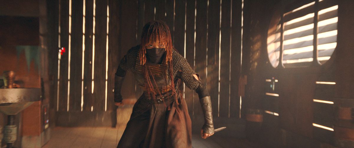 Mae (Amandla Stenberg) in The Acolyte.  She stands in a fighting stance in a wooden slatted building, holding a small dagger in one hand.  The bottom of her face is covered with a cloth mask.
