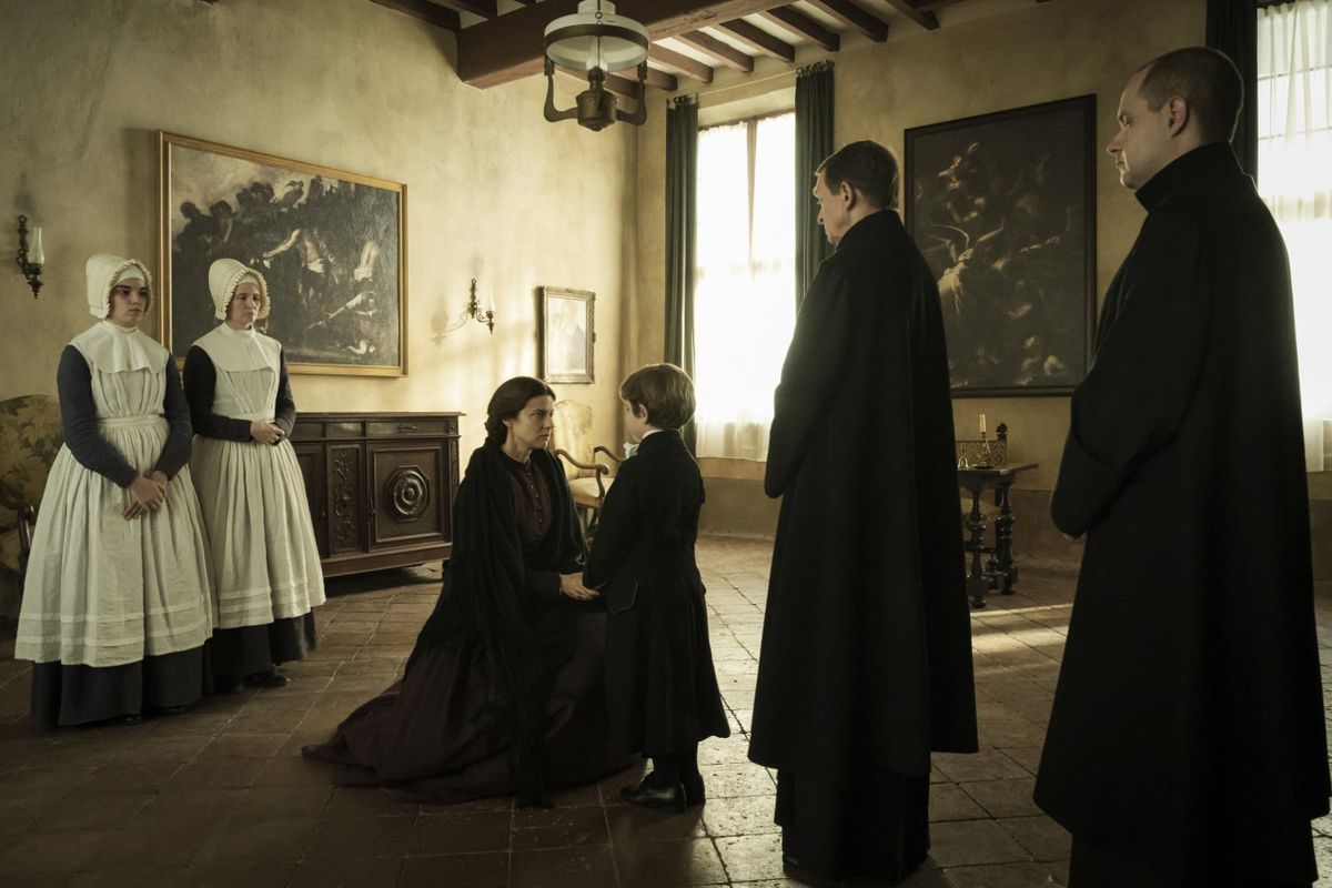 A woman kneels before a young boy in a room surrounded by maids and clerics in Kidnapped; The Abduction of Edgardo Mortara.