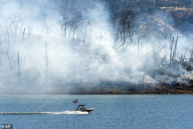 A boat crosses Lake Oroville with a smoldering hillside behind it as the Thompson Fire rages in Oroville, California, on Wednesday, July 3, 2024