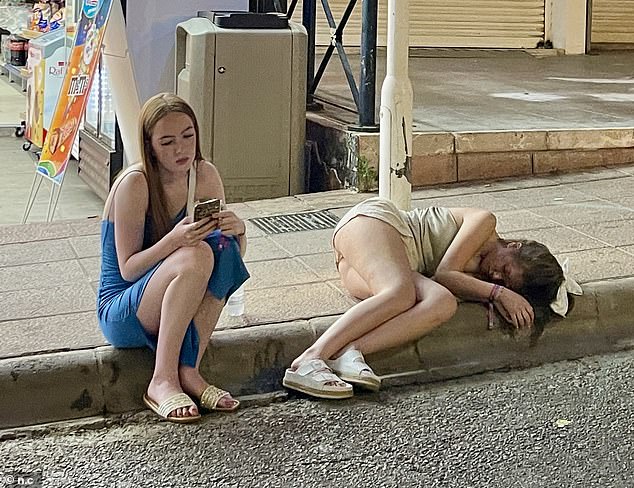 A vacationer takes a nap on the sidewalk while her friend sits next to her after a night out