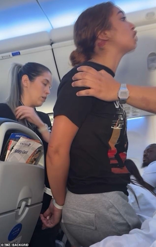 The video cuts to the woman, now standing in the aisle with her hands tied behind her back, yelling at everyone not to fly United.