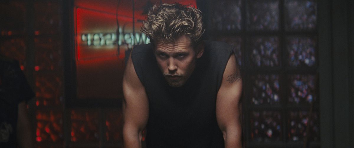 Austin Butler, with tousled hair and a black sleeveless top, leans forward moodily in The Bikeriders