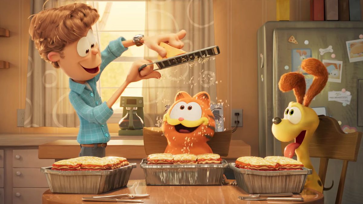 Jon Arbuckle grates Parmesan cheese over Garfield's lasagna as Odie looks on in a still from The Garfield Movie