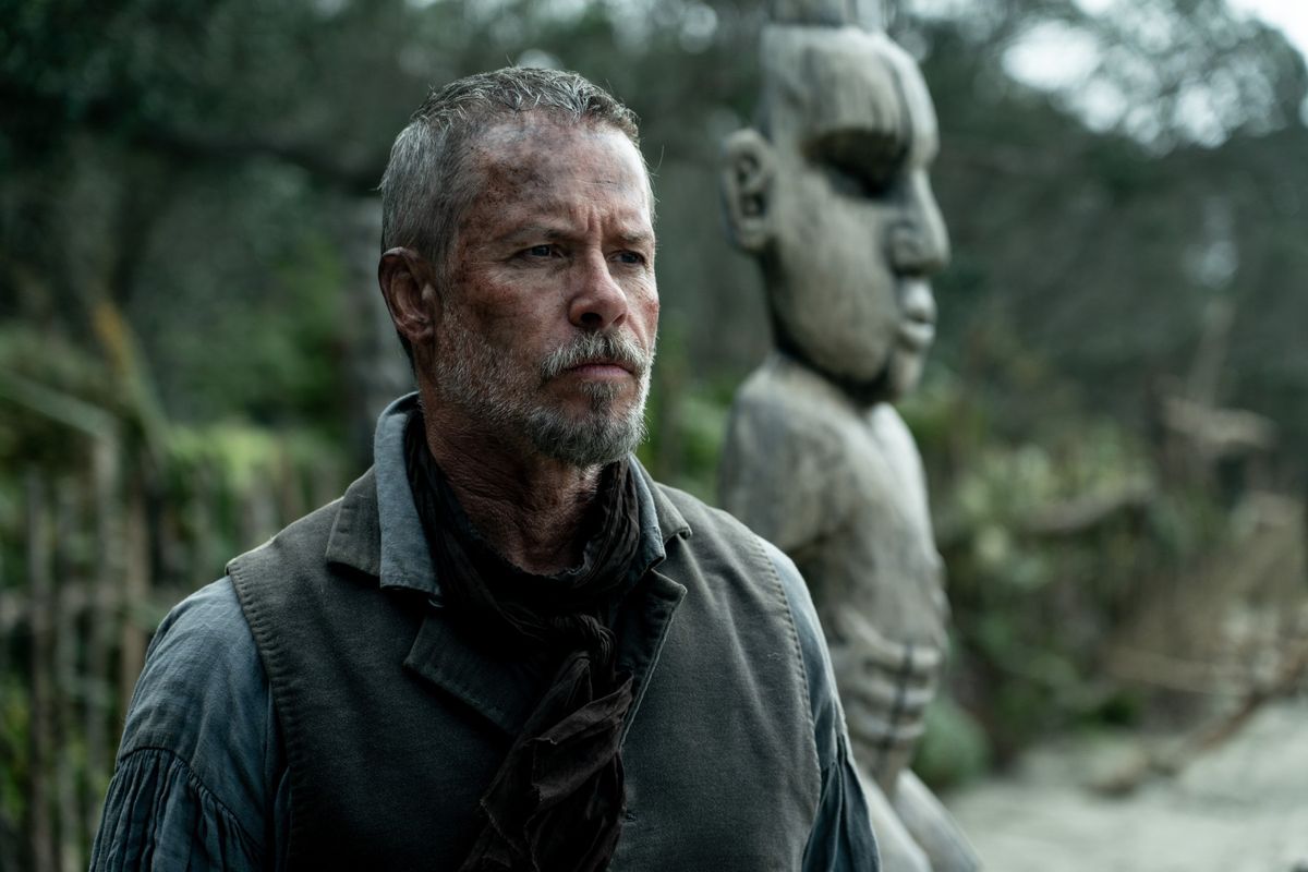 A stern-looking man with a beard and bruises on his face stares off into the distance, with a large wooden totem behind him in The Convert.