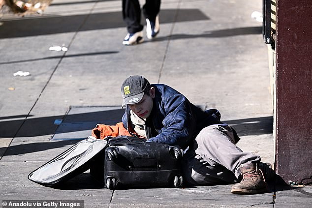 Homeless people gather downtown, pushing their belongings into shopping carts or sitting on sports bags. The photo shows a homeless man on the sidewalk