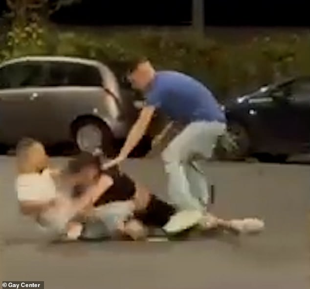 The attacker in blue and the attacker in white can be seen holding a belt as he whips the gay man in black after he is tackled to the ground