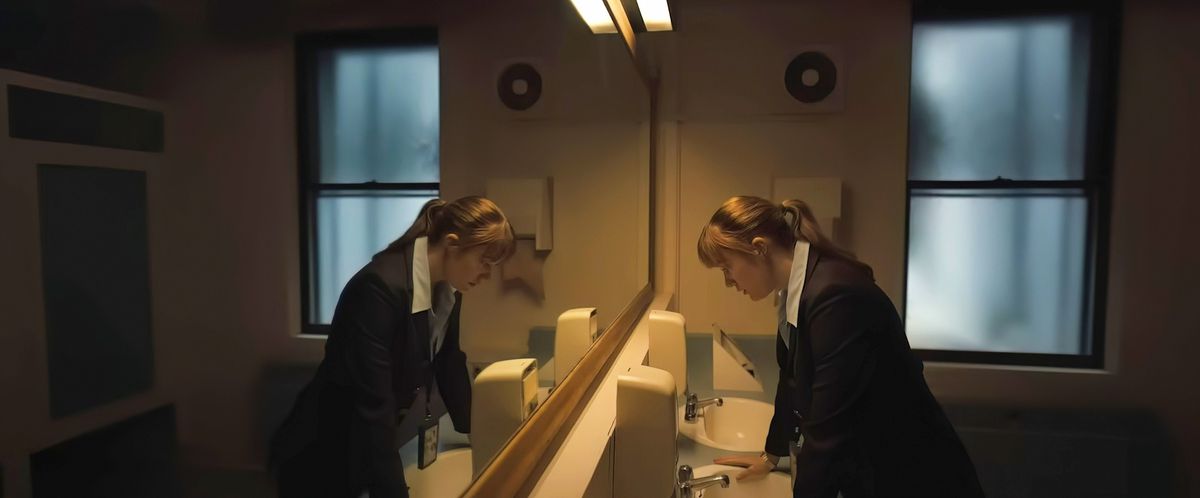 FBI agent Lee Harker (Maika Monroe) stands in a large bathroom by a row of sinks and stares down, reflected in a wall-length mirror, in Oz Perkins' Longlegs