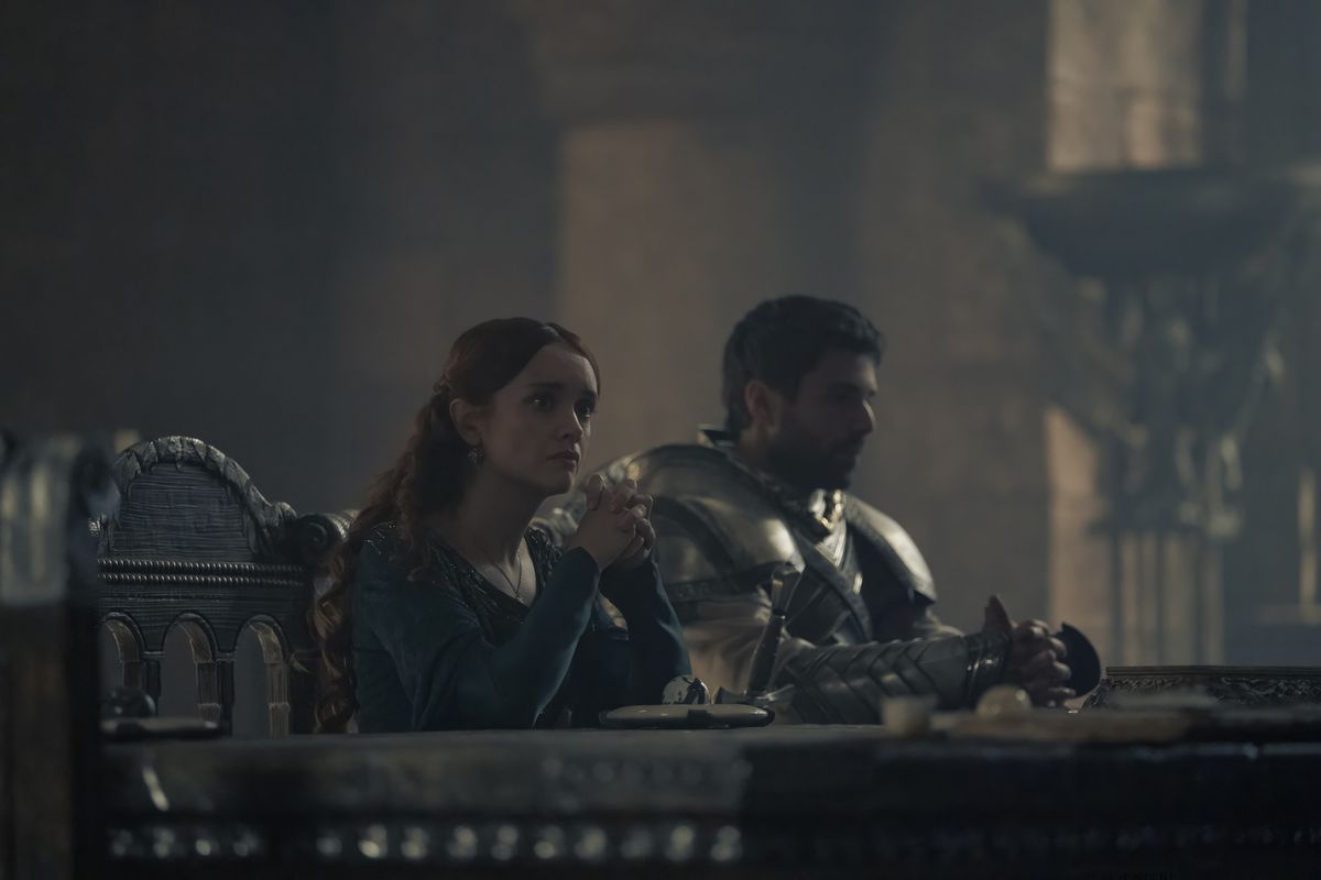 Alicent (Olivia Cooke) sits at the small council table with her hands folded and looks worried, with Criston Cole (Fabien Frankel) in the background