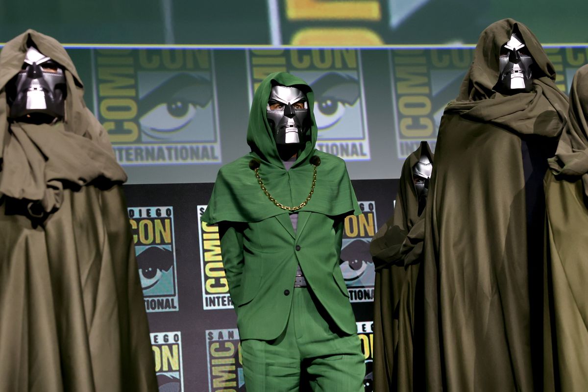 Robert Downey Jr. in a green hooded suit and gold chain (very Doctor Doom) and wearing a Doctor Doom mask onstage during the Marvel Studios Panel at SDCC 2024. 