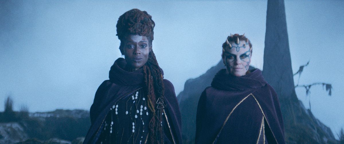 (L-R): Mother Aniseya (Jodie Turner-Smith) and Koril (Margarita Levieva) in The Acolyte. They stand side by side, slightly smiling, in elaborate robes on a barren cliffside.