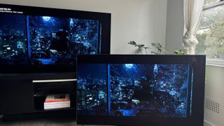 The LG G4 OLED TV on a TV stand, with the LG G3 OLED next to it on the floor, both showing the same scene of a man being thrown out of a glass window, in the movie Watchmen