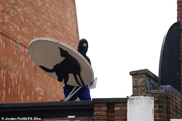 A man wearing a hood and a mask holding the satellite dish while standing on the roof of the building