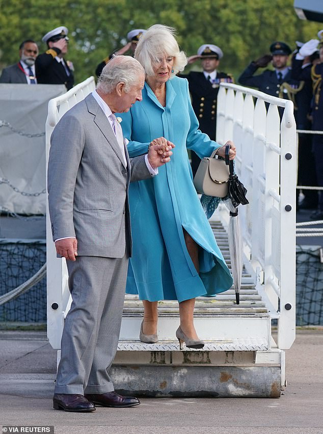 The King helps Camilla, dressed in a long blue coat and carrying a beige handbag, to disembark from the flight deck of HMS Iron Duke in Bordeaux during the couple's state visit to France, September 2023