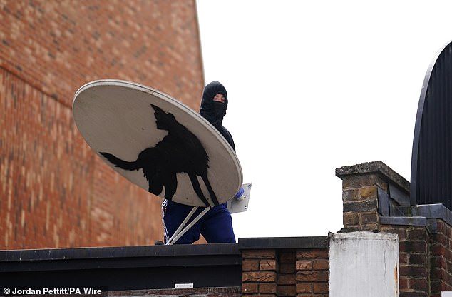 A masked man in a hood is seen holding a satellite dish featuring Banksy's wolf mural before the work disappeared