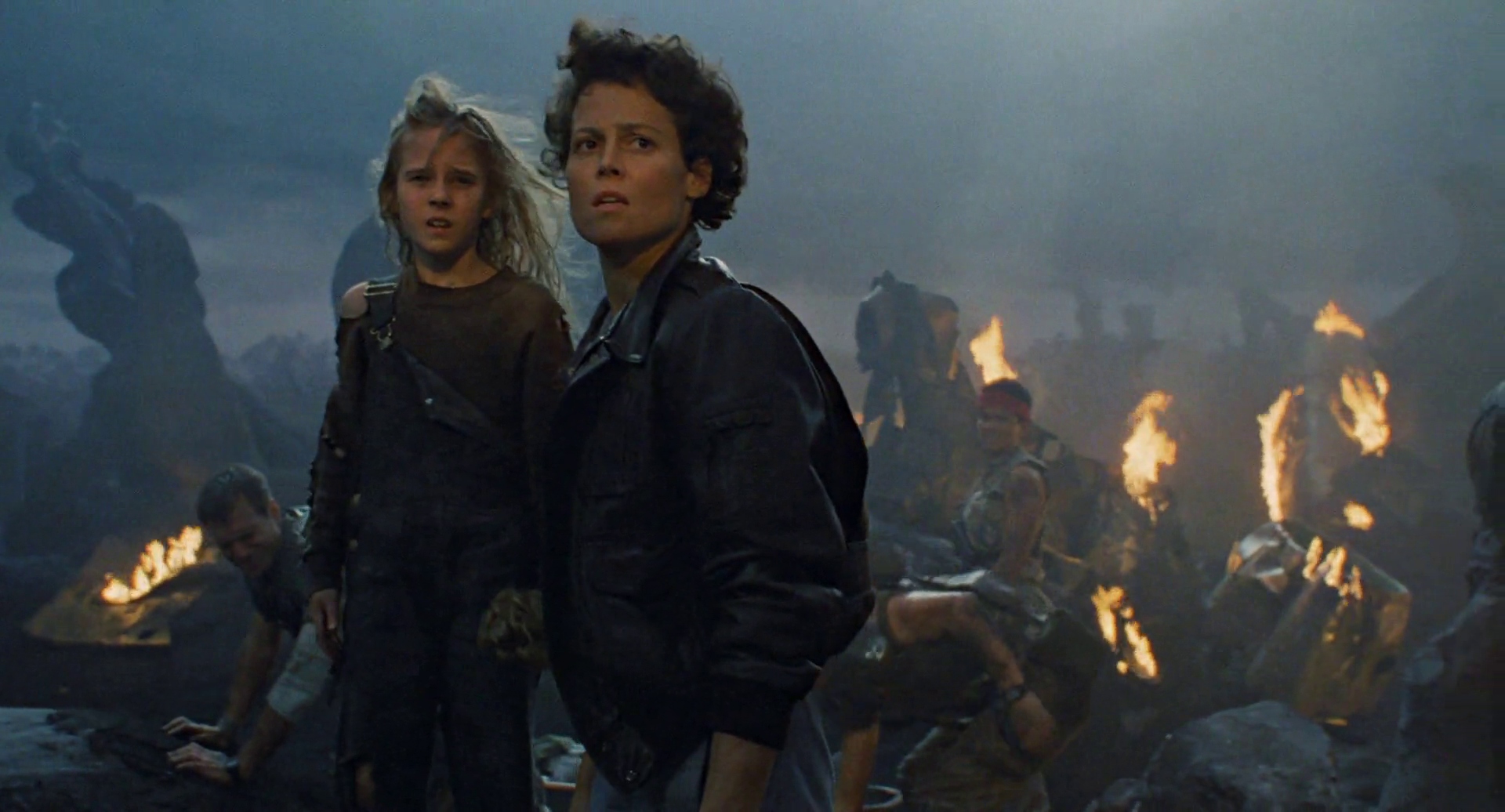 (L-R) Carrie Henn and Sigourney Weaver as Newt and Ellen Ripley, surrounded by wreckage, fire, and wounded Colonial Marines in Aliens.