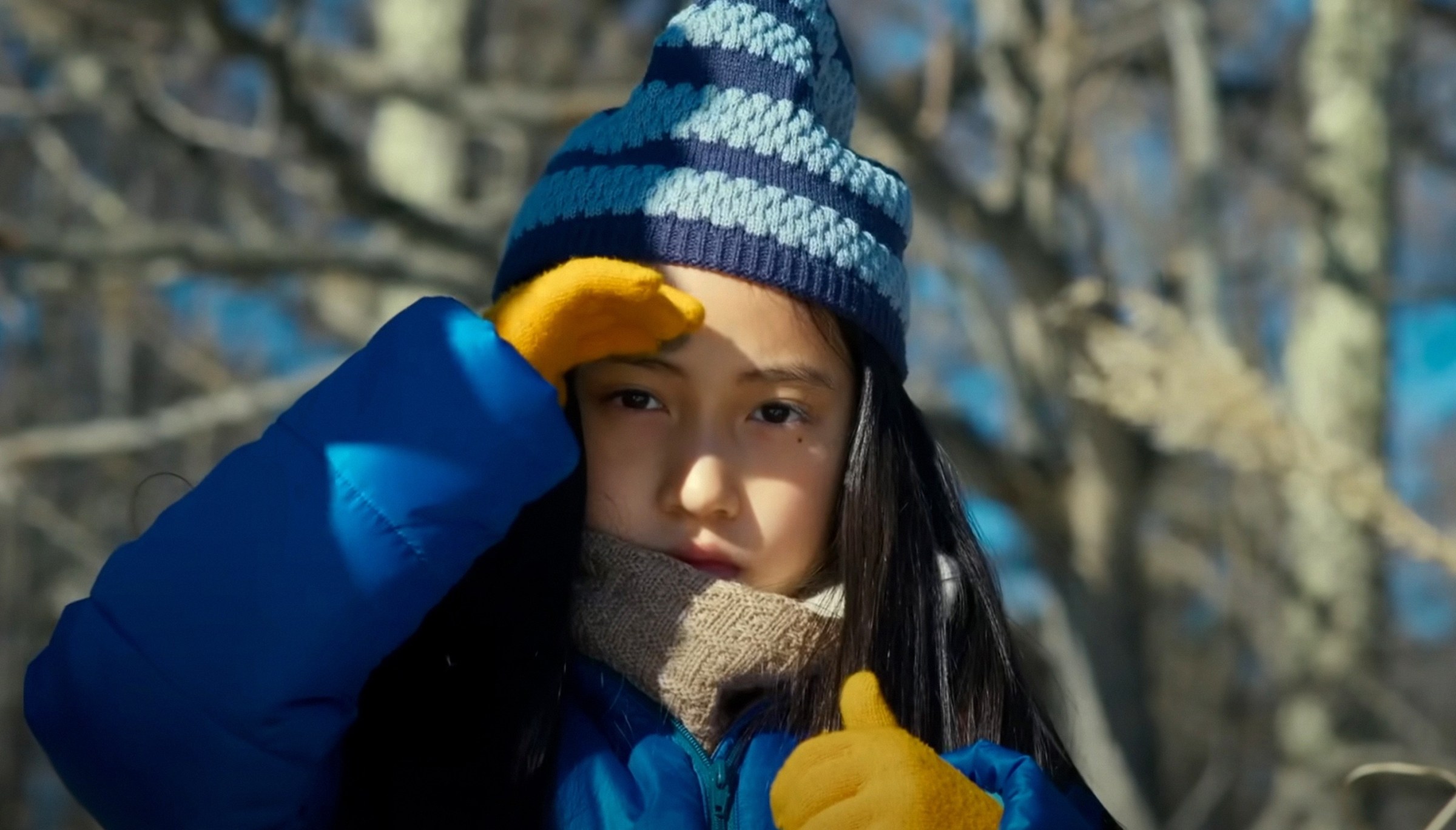 Hana (Ryo Nishikawa), a young Japanese girl in a thick coat and knitted hat, holds her hand over her eyes and looks doubtfully into the camera in Ryûsuke Hamaguchi's Evil Does Not Exist