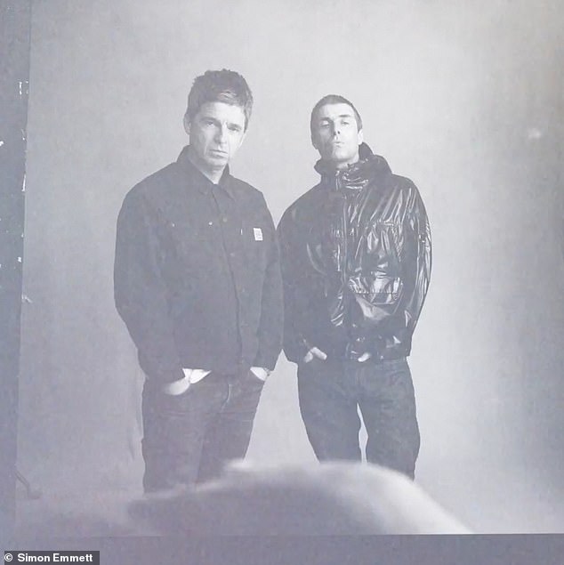 In this photo, Liam is standing behind Noel, leaning back slightly with his hands in his pockets