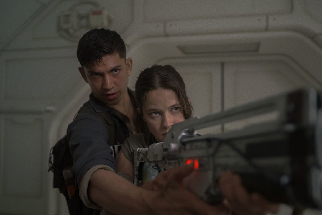 A man stands over a woman's shoulder as she lurks with a futuristic rifle in Alien: Romulus.A man stands over a woman's shoulder as she lurks with a futuristic rifle in Alien: Romulus.