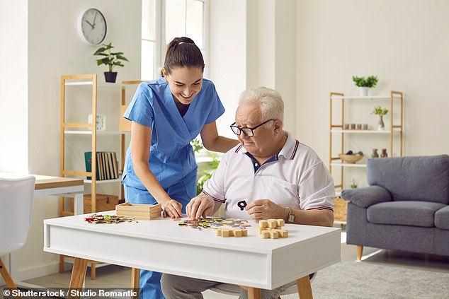 Smiling young female social worker and old man with Alzheimer's play with a jigsaw puzzle in a nursing home.