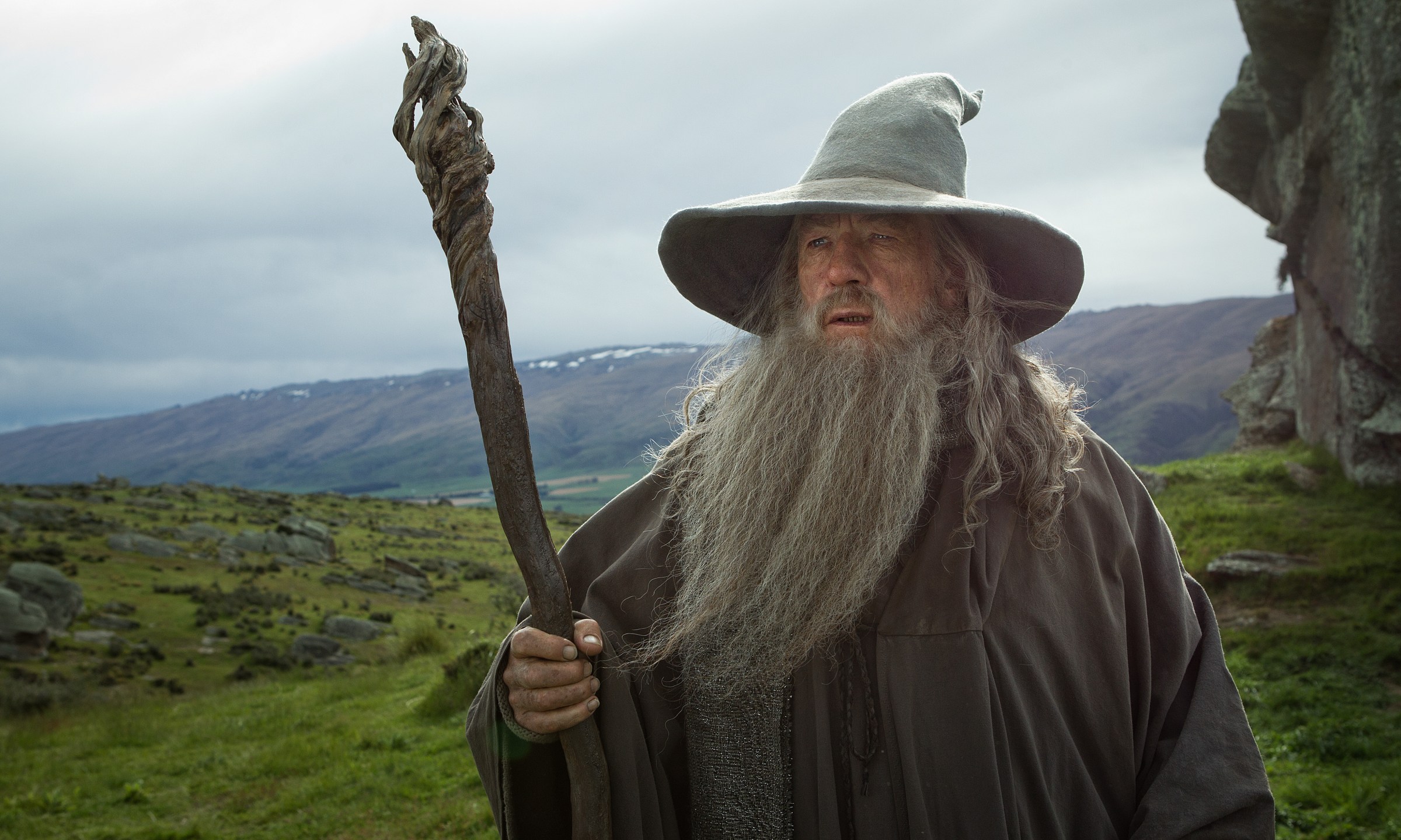 Gandalf the Grey holds his staff with a green, hilly field behind him in The Hobbit: An Unexpected Journey