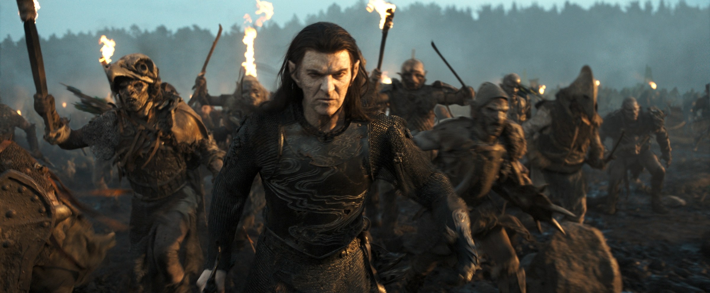 Sam Hazeldine as Adar the corrupt elf, the ancient leader of an army of orcs in The Lord of the Rings: The Rings of Power. He walks through a battlefield, orcs gathering behind him, weapons drawn.