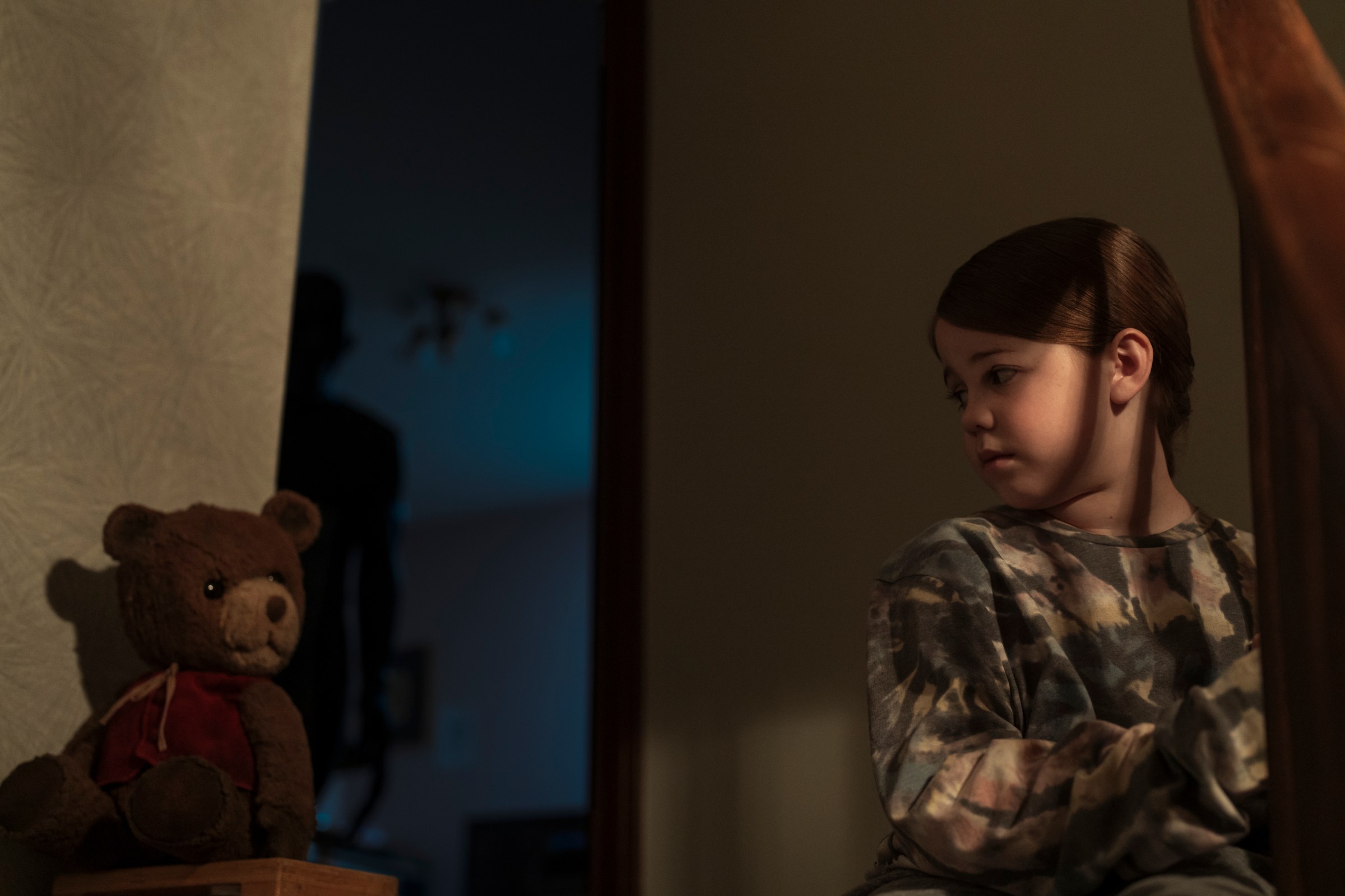 A young girl played by Pyper Braun sits at the top of the stairs next to a teddy bear as an ominous shadow figure lurks behind her in Imaginary