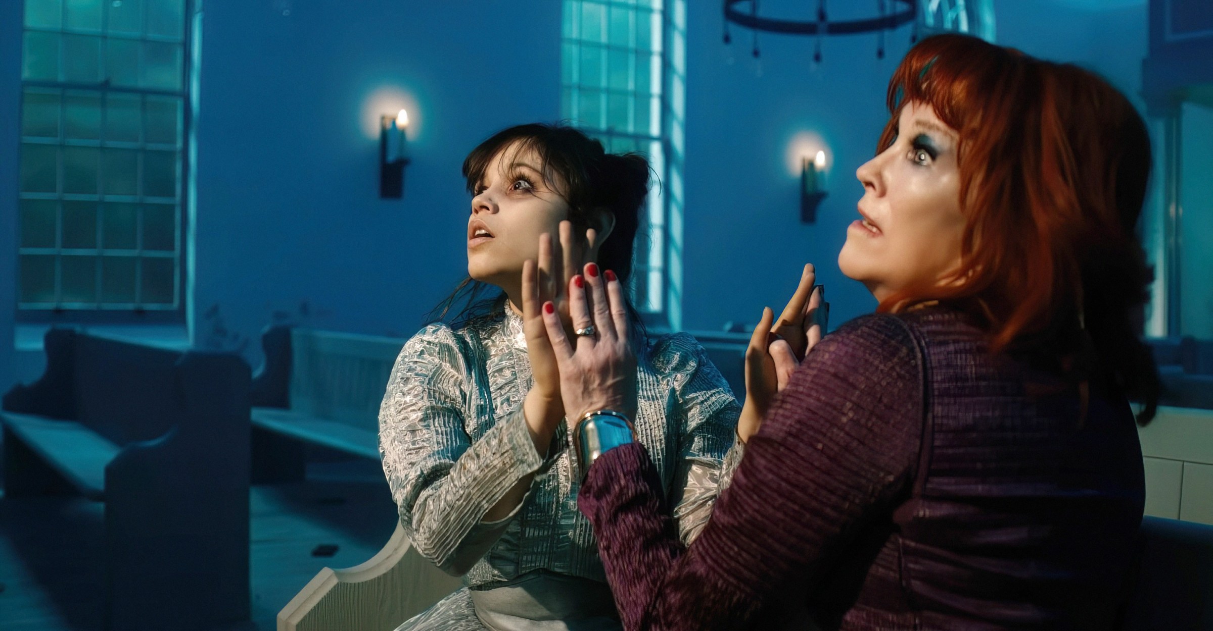 Astrid and Delia Deetz (Jenna Ortega, Catherine O'Hara) sit in a pew, holding their hands up and pressing their palms together, looking up and to the side at something off-screen in Beetlejuice Beetlejuice