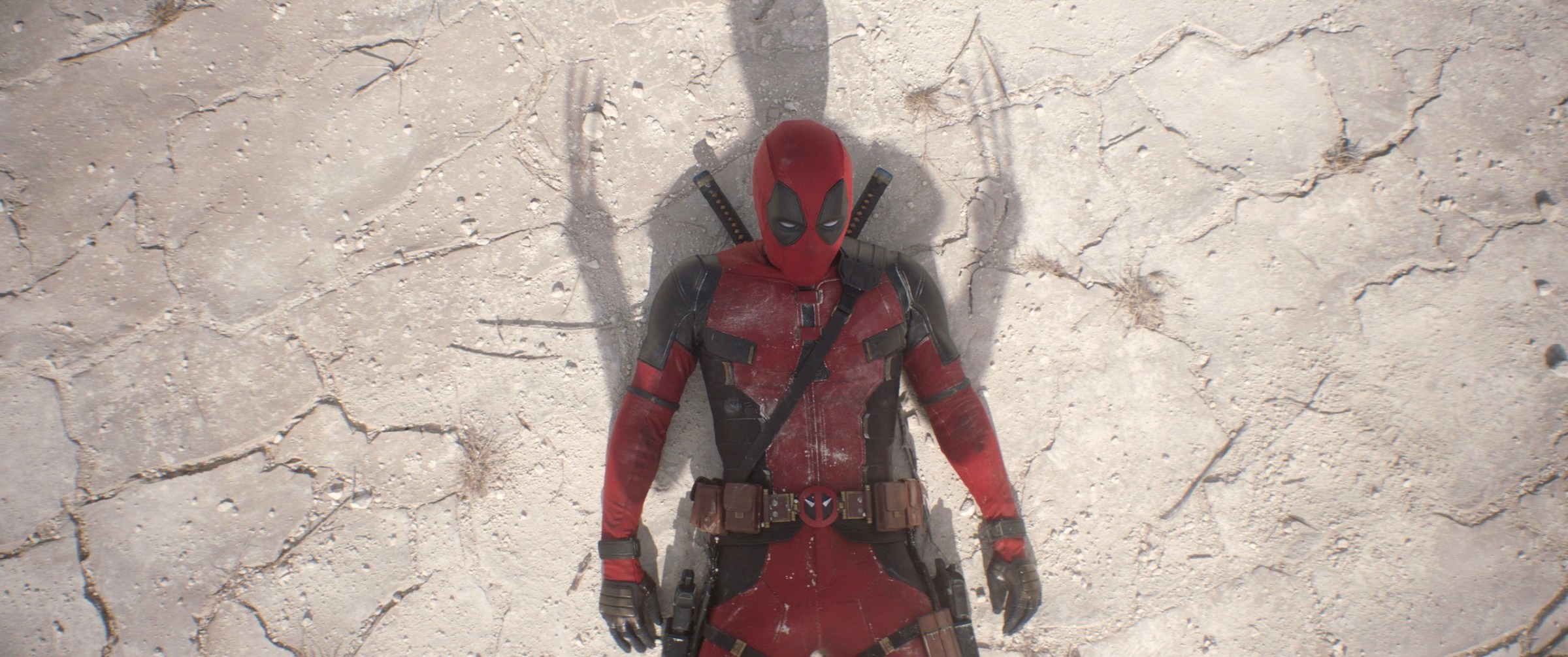 Deadpool (Ryan Reynolds) lies on his back in the sand with Wolverine's shadow over him, claws out, in Deadpool & Wolverine