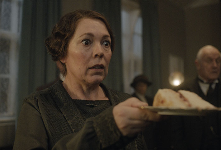 Olivia Colman, surprised, holds a piece of cake on a plate in Wicked Little Letters