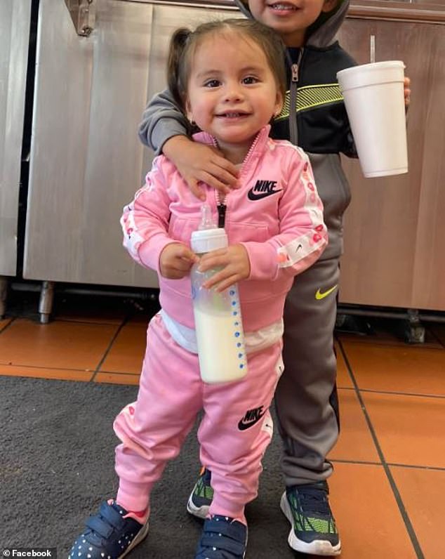 The child in the photo with her older brother is holding a bottle and smiling