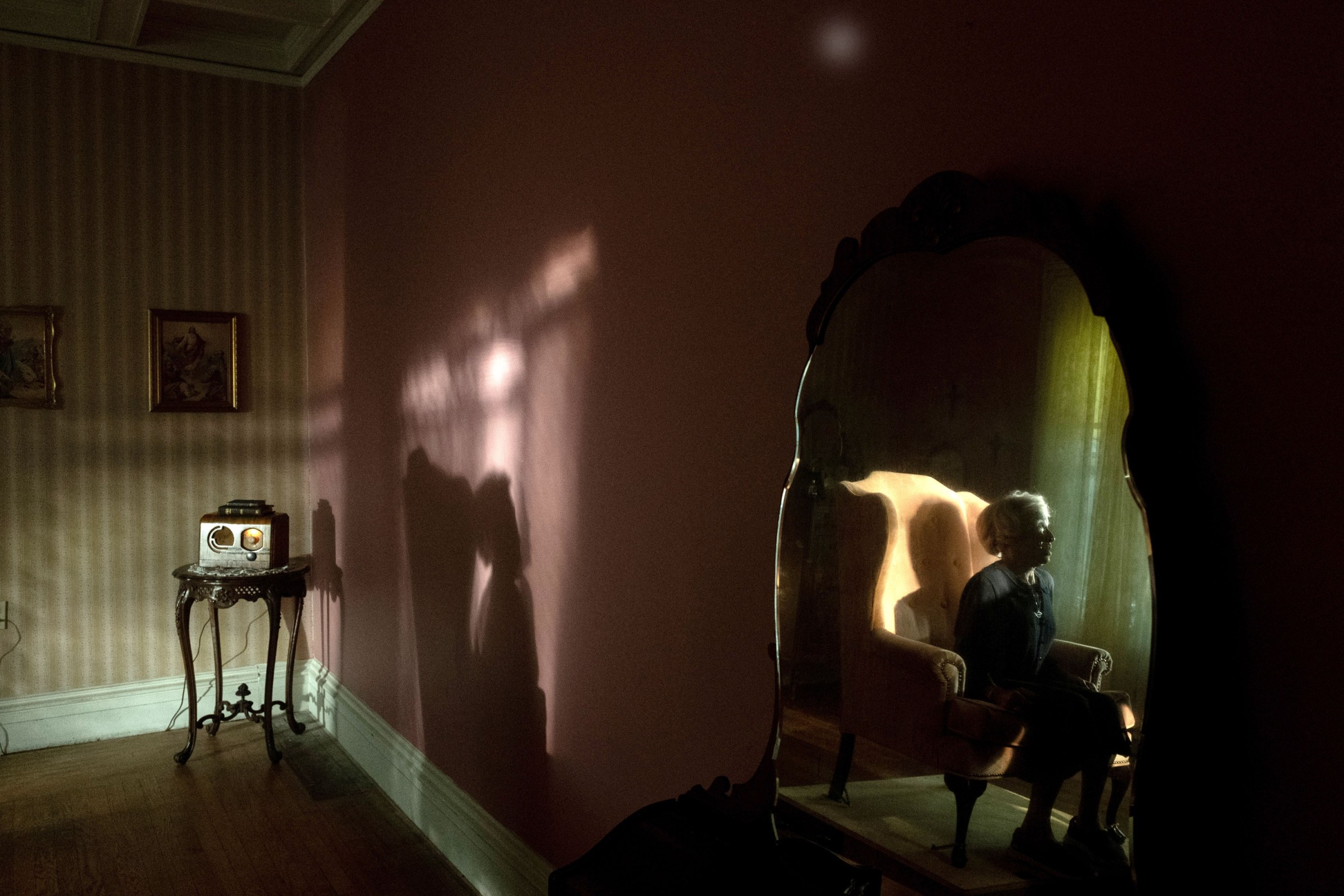 A silhouetted figure sits in what appears to be a hole in a wall in a dimly lit room in a dreamy scene from Sam and Max Eggers' horror film The Front Room