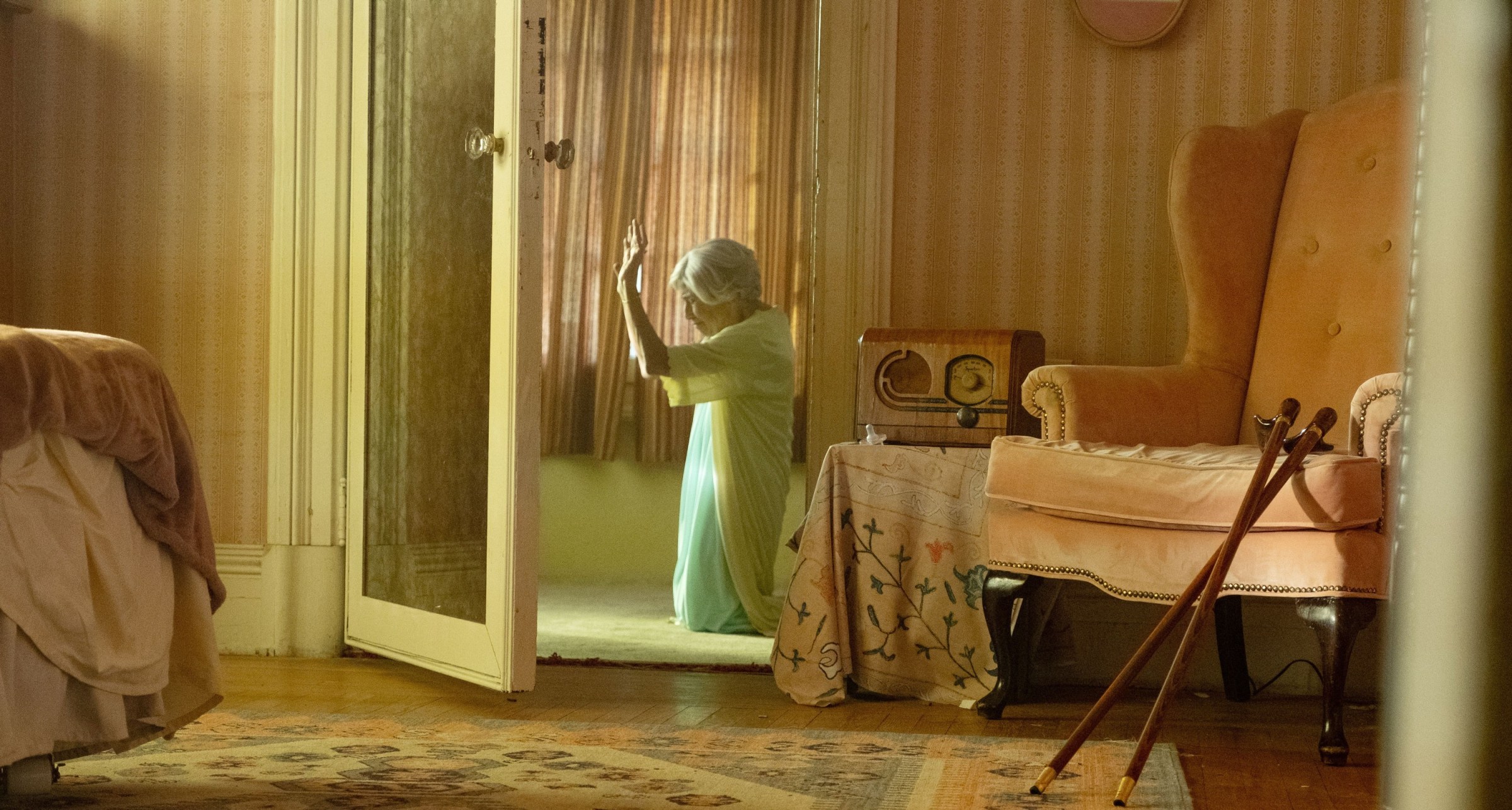 Solange (Kathryn Hunter), the abusive mother-in-law who plays the villain in Sam and Max Eggers' A24 horror film The Front Room, kneels in prayer in a sunbeam, in a shot captured through a door and an adjacent room.