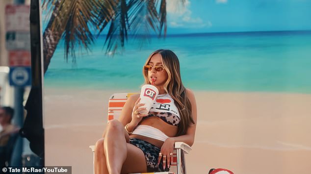 The video starts off innocently enough with McRae dressed in a beach outfit, sitting on a lounge chair in a tropical location, sipping on a soda