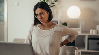 A woman bends over, holding her back with one arm and looking at a laptop that is on a surface