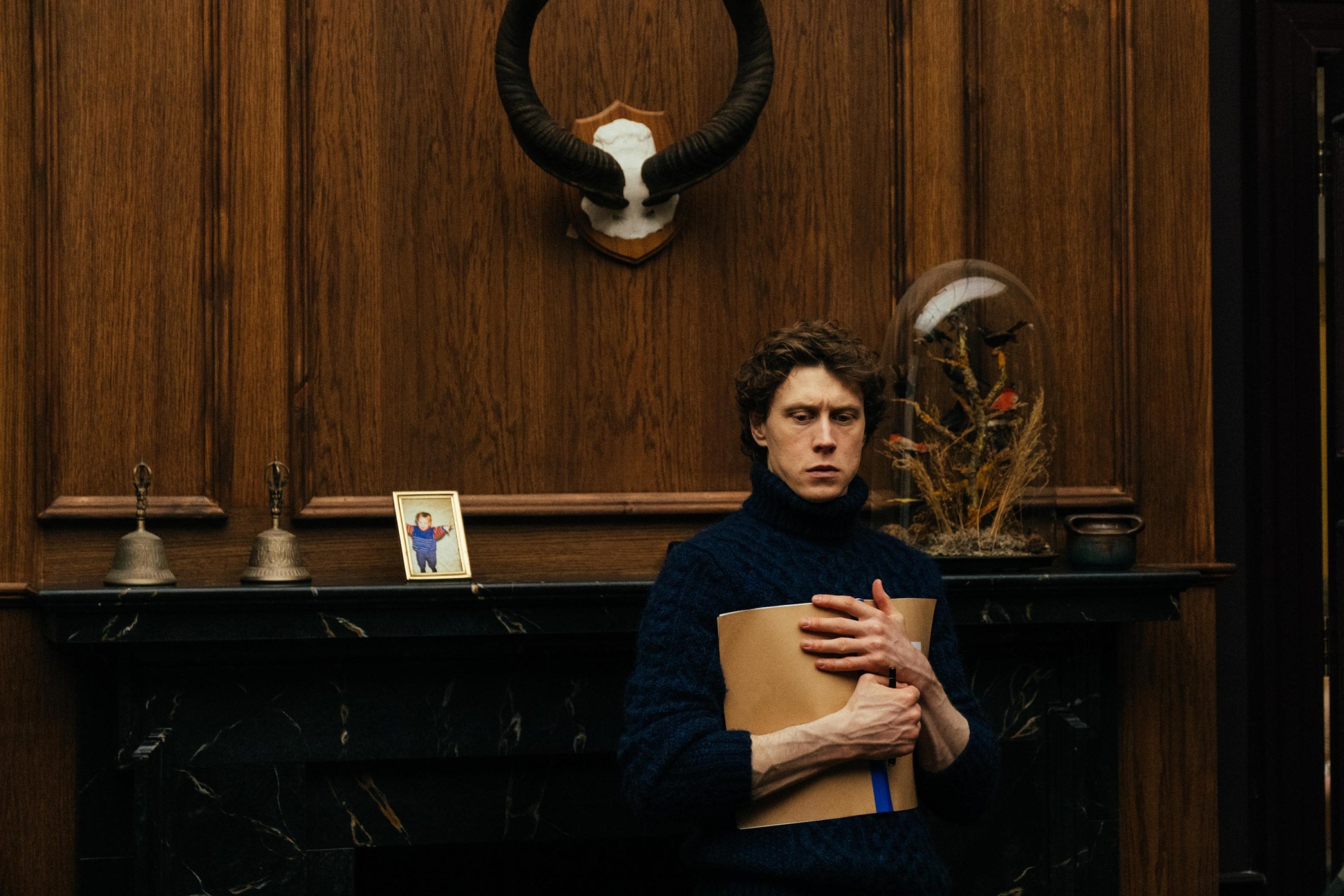 George MacKay, clutching a brown paper folder to his chest, stands before a wide fireplace surrounded by dark wood paneling in The End