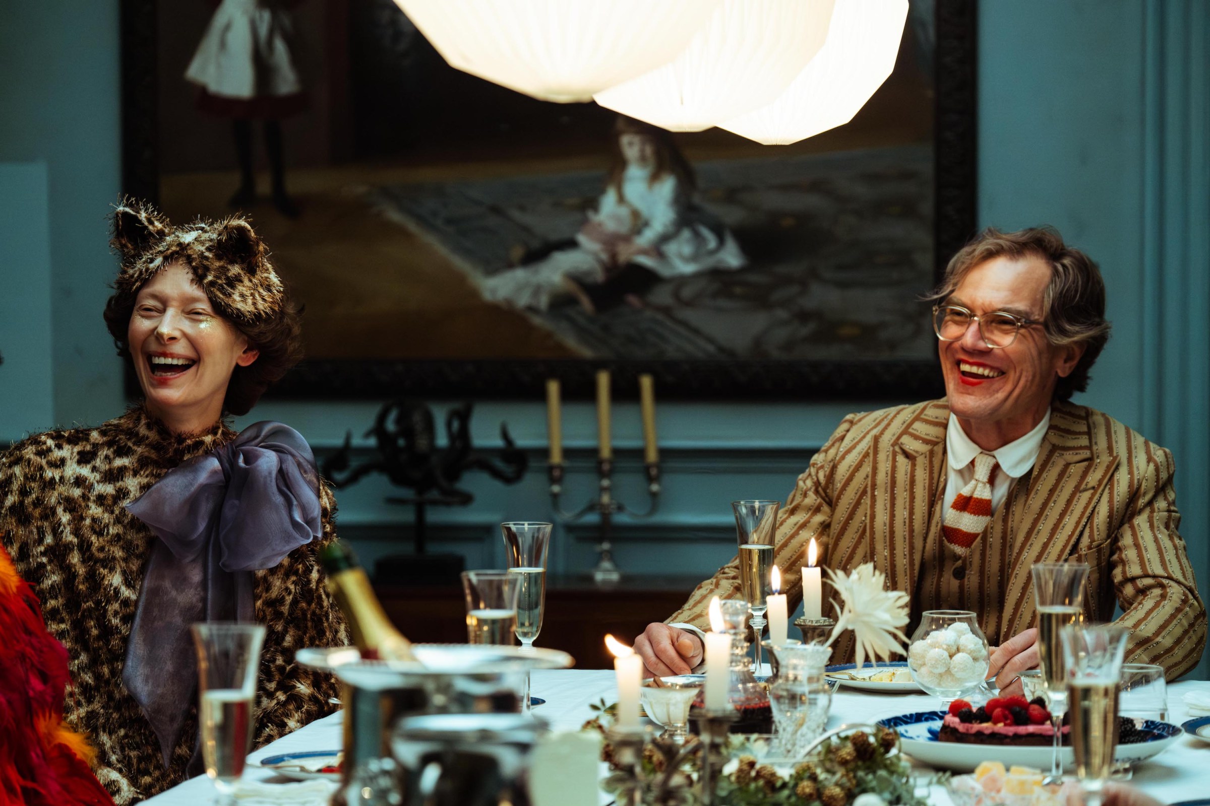 Tilda Swinton (wearing a leopard fur hat, complete with cat ears, and a leopard dress with an immense blue bow) and Michael Shannon (in a brown, vertically striped suit with wide lapels) sit together, laughing, at a lavishly set table in The End