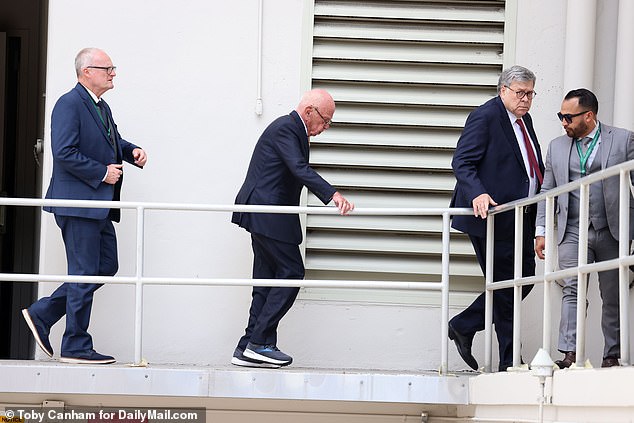 Murdoch grabs the railing for support as he leaves the hearing, with former US Attorney General Bill Barr, who is acting as one of his lawyers, standing before him.