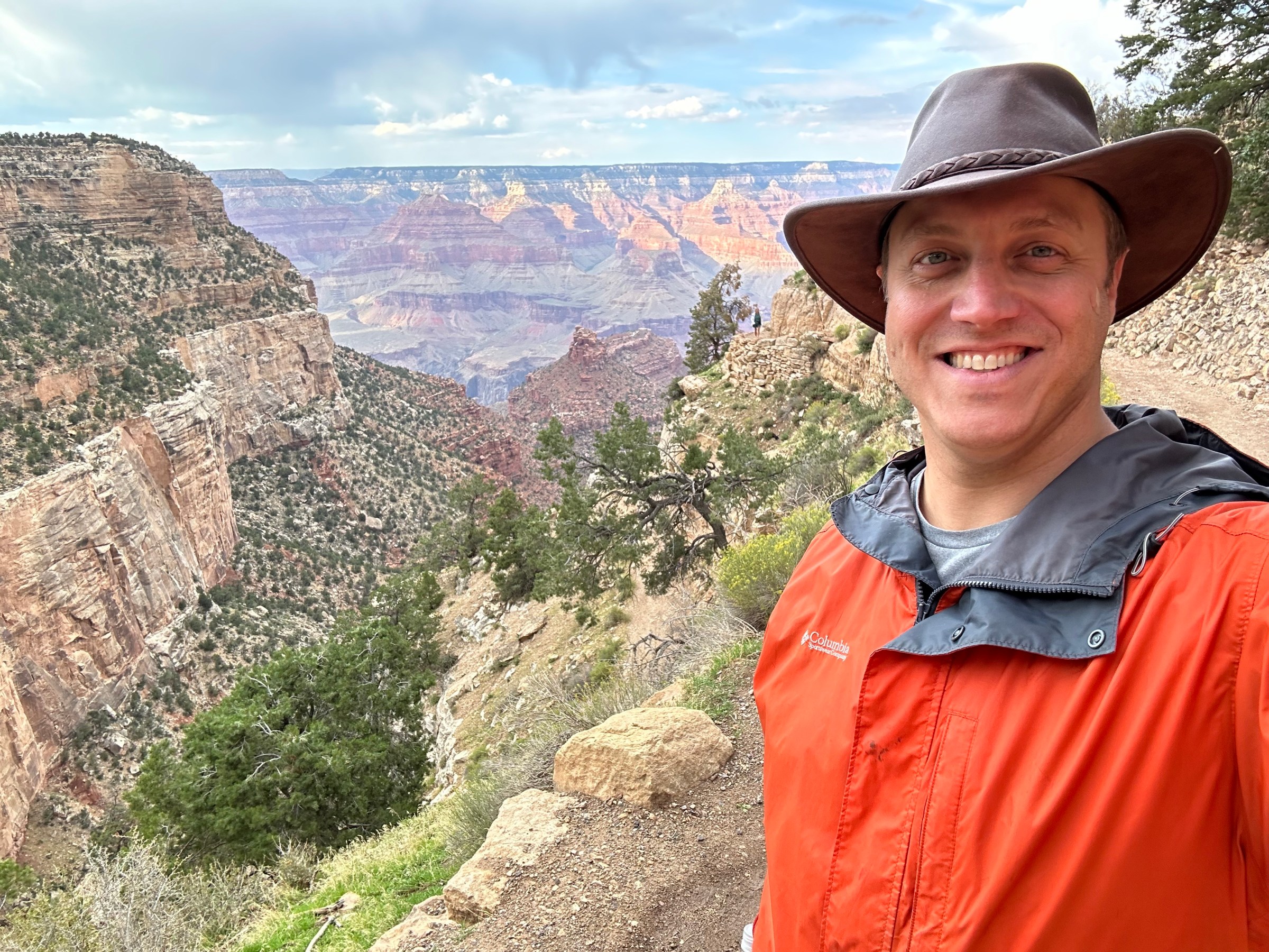 A man wearing a leather hat and an orange raincoat stands on the edge of a gorge.