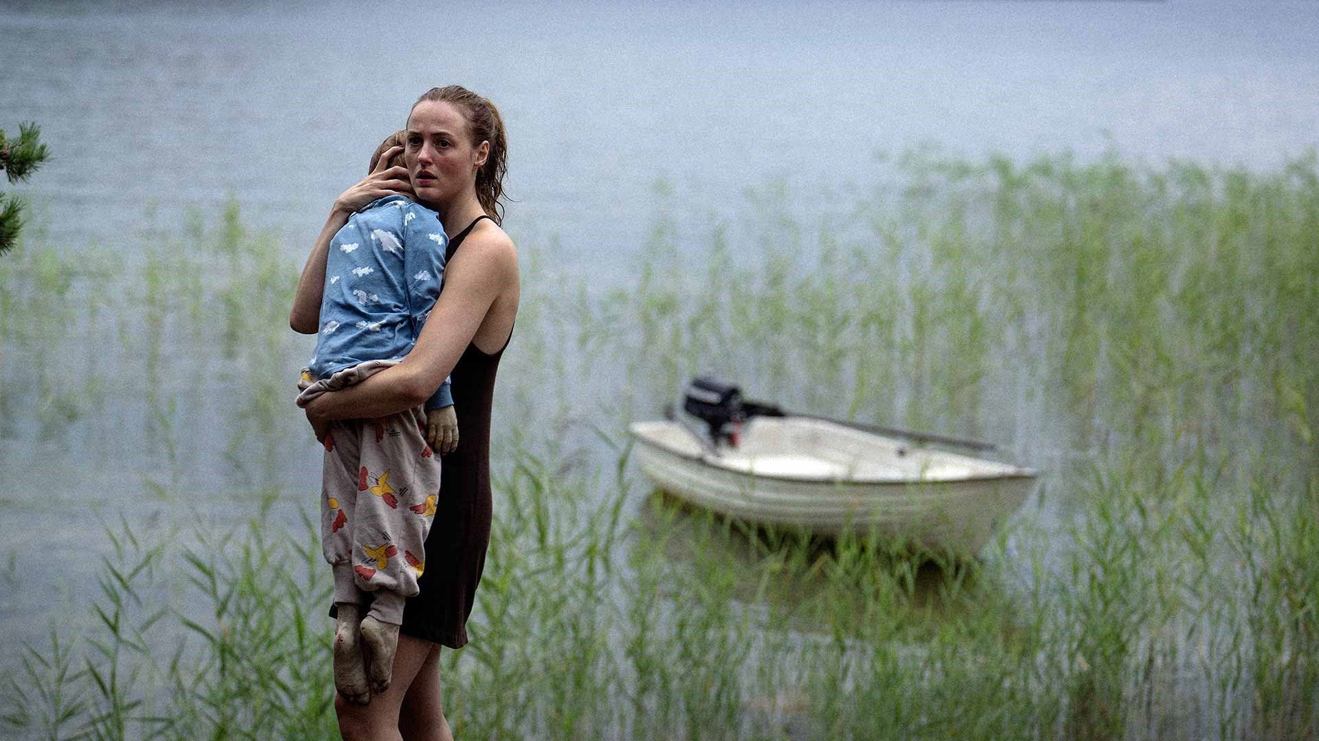 A woman holds the limp body of a child in her arms next to a boat, surrounded by a marsh of reeds in Handling the Undead.