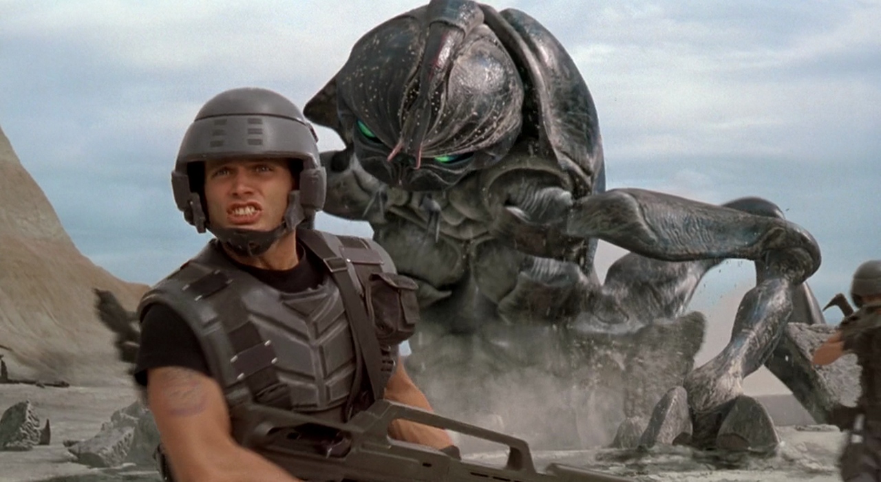 A man in soldier armor stands in front of a giant insect in Starship Troopers.