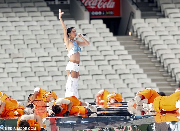 Katy stands at the front singing into the microphone, dressed in her metallic blue crop top and white shorts with attached, wide leg sleeves, continuing the futuristic theme